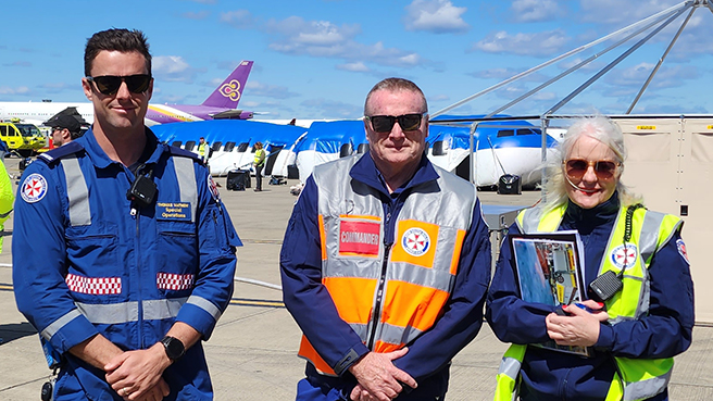 A photo of Special Operations Team Paramedic Thomas Mathew, Chaplain Jane Mahon and Inspector Geoff Senior on the tarmac at Sydney Kingsford Smith Airport taking part in Exercise Galavanise - a plane crash emergency simulation - on 2 November 2022. Behind them is a full size blow-up model of a crashed airplane.