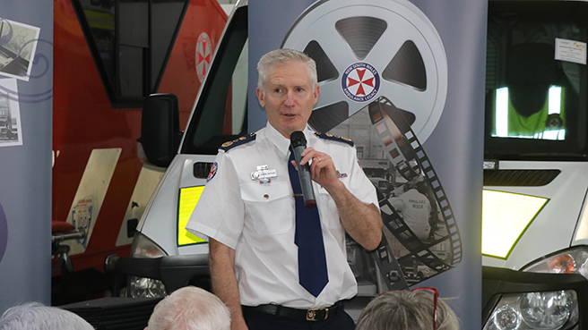 A photo of NSW Ambulance Chief Executive Dr Dominic Morgan addresses attendees at an Ambulance Legacy event  at Temora Ambulance Museum on 22 October 2022. Dr Morgan is standing in front of pull up banners and a NSW Ambulance vehicle.